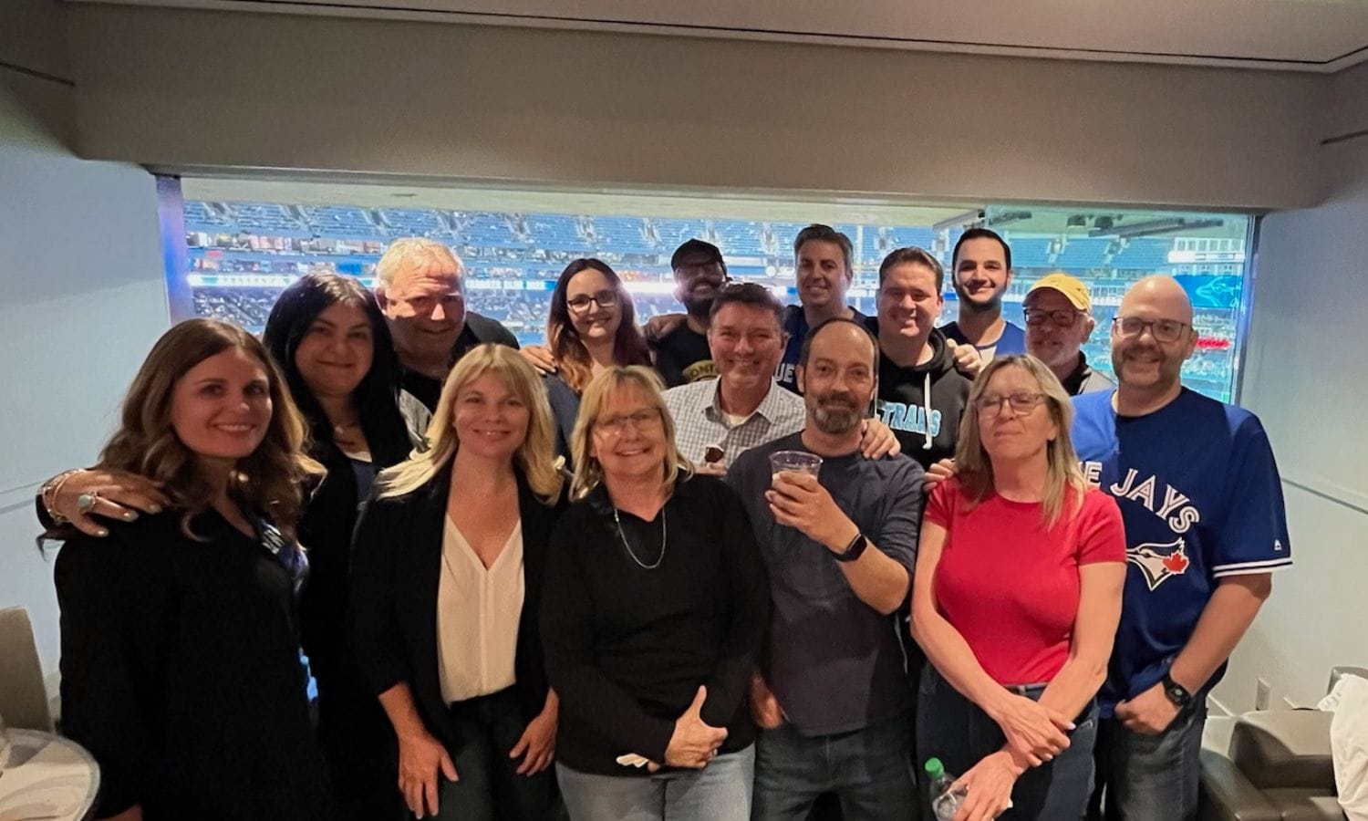 A group of fourteen people, including men and women, are posing for a photo in what appears to be a stadium suite. Some are smiling at the camera, while others are holding drinks. The image captures a casual, cheerful atmosphere at a sporting event.