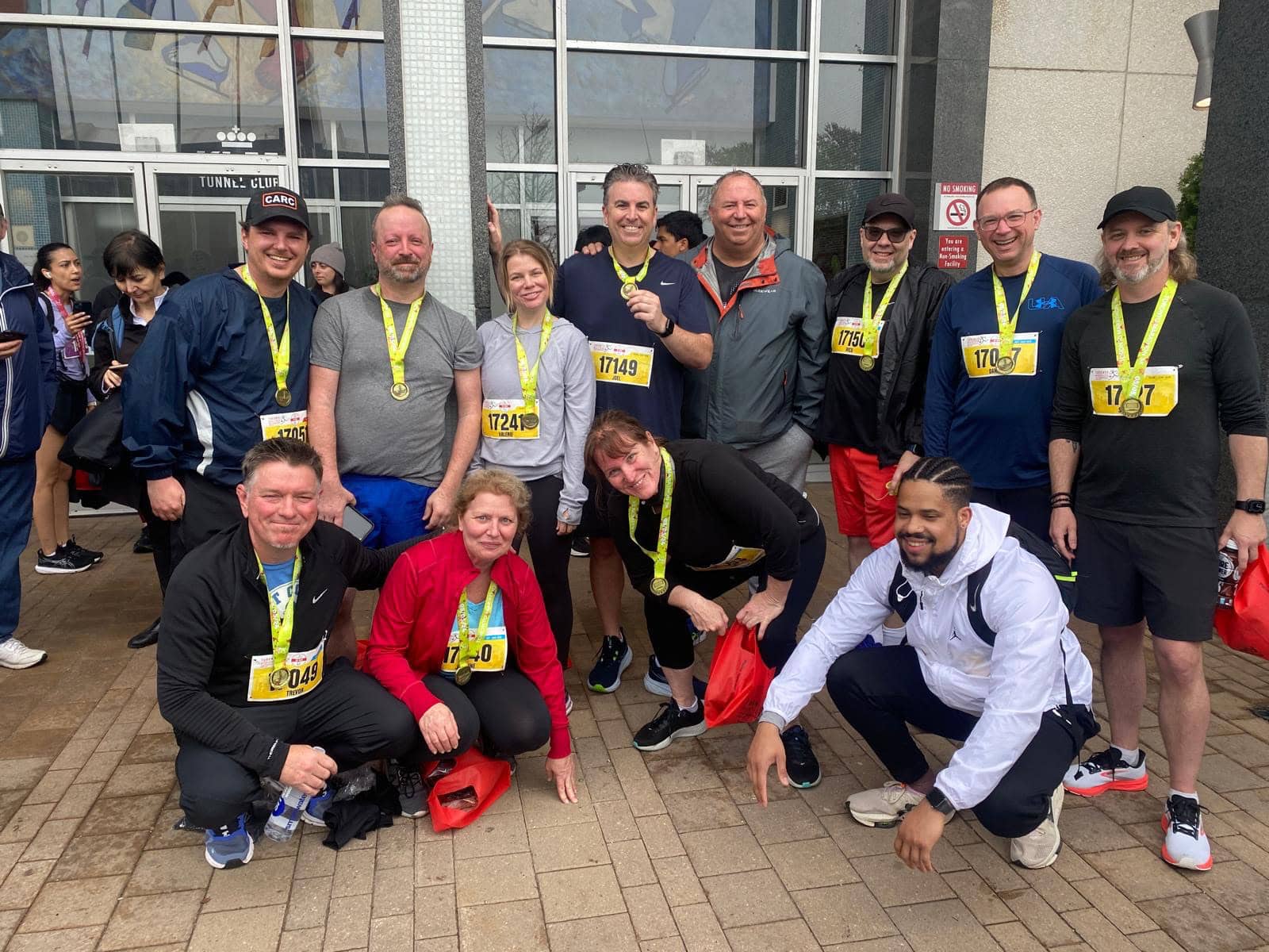 A group of people posing for a photo after a race, wearing medals and bib numbers. They are smiling and dressed in running gear. Some are standing, while others are kneeling. The background shows a building with glass windows and other participants.