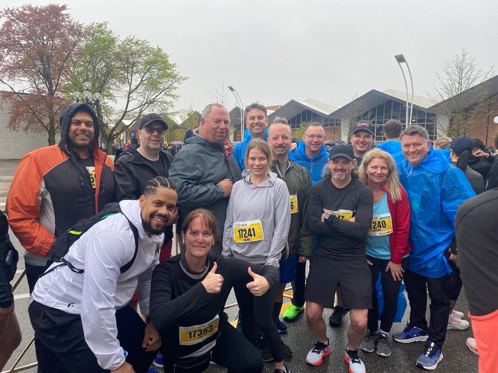 A group of people in athletic gear pose and smile at a running event. All wear race bibs, and some have jackets or hats. It's overcast, with trees and modern buildings in the background. One woman in the front gives a thumbs-up.